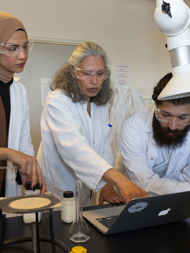 To elever og en gymnasielærer i kitler kigger på en computer i et naturfagslokale 