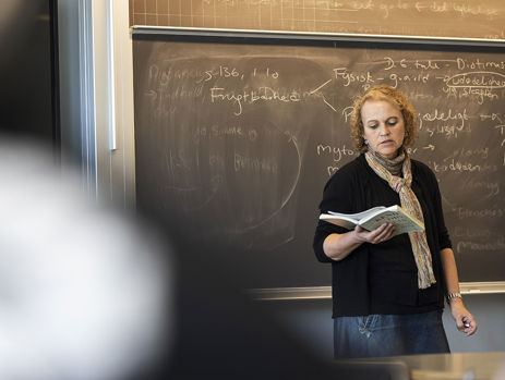 Gymnasielærer læser højt fra bog foran en tavle i et klasselokale 