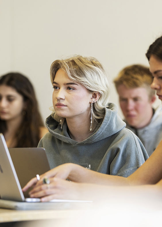 Elever sidder i et klasselokale. En elev rækker hånden op, mens andre arbejder og ser koncentreret ud